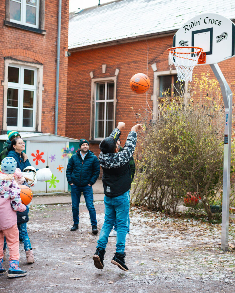 Ein Kind wirft den Basketball auf den neuen, von Lindner Bau fundierten Korb in Lugau. Im Hintergrund sind Erwachsene des Bauunternehmens und der Ridin' Crocs zu sehen, die das Bauwesen-Projekt unterstützen.