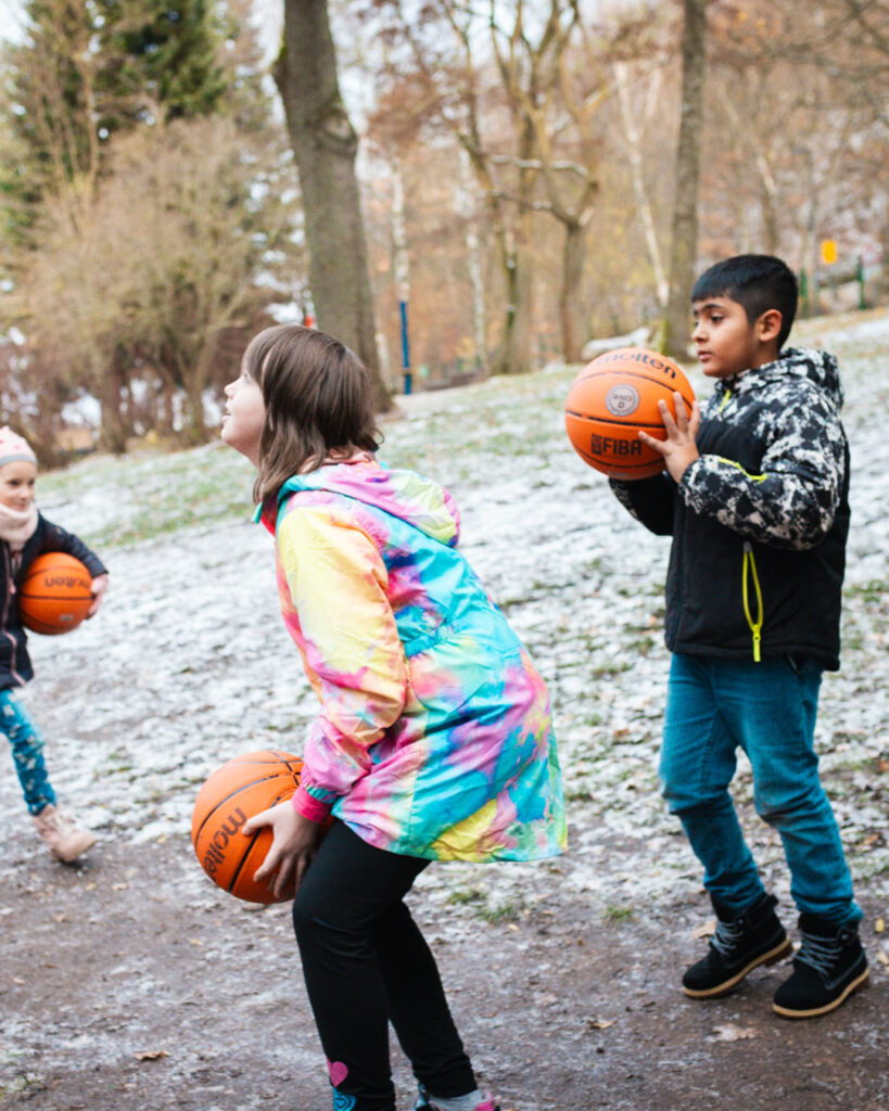 Kinder spielen mit Basketbällen auf dem Gelände der Schule in Lugau mit dem neuen Korb. Das Bild hebt die Freude über die Spende und das Engagement von Lindner Bau im Bereich Sport und Sanierung hervor.