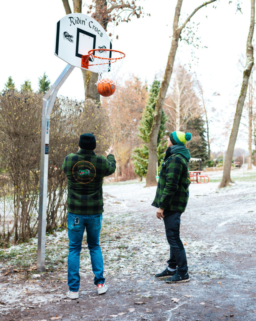 Zwei Erwachsene werfen den Ball auf den "Ridin' Crocs" Basketballkorb im Freien. Das stabile Fundament des Korbes wurde von Lindner Bau als Teil der Schul-Challenge in Sachsen gesetzt.