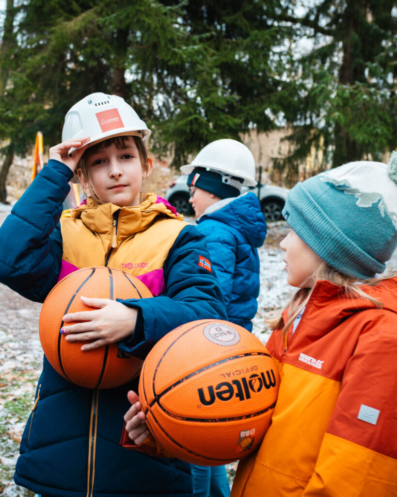 Kinder in Winterkleidung mit Basketbällen und Bauhelmen am neuen Korb in Lugau. Zeigt die Übergabe des durch Lindner Bau fundierten Korbes nach der erfolgreichen Schul-Challenge.