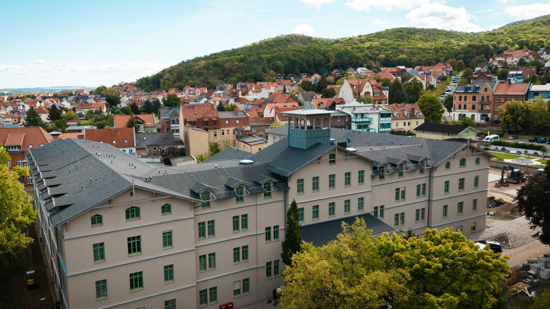 Luftaufnahme eines großen U-förmigen Gebäudes mit dunklem Dach und grünen Fensterrahmen in einer städtischen Umgebung, umgeben von kleineren Häusern und Bäumen.