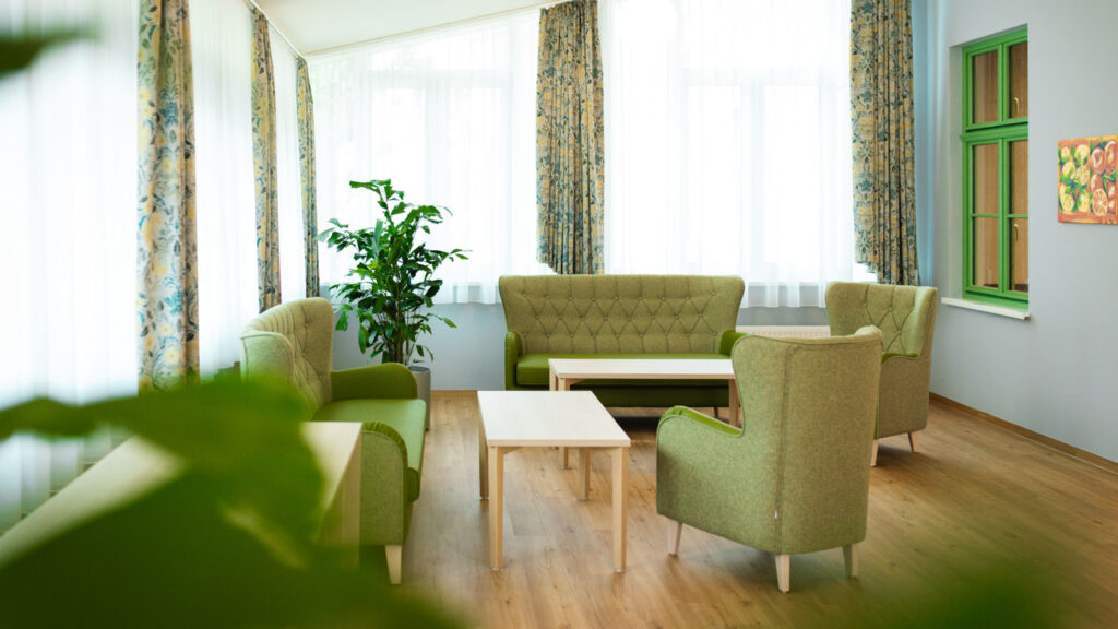 Heller Gemeinschaftsraum mit vier bequemen, hellgrünen Polstermöbeln und zwei schlichten Holztischen. Große Fensterfronten sorgen für viel Tageslicht.