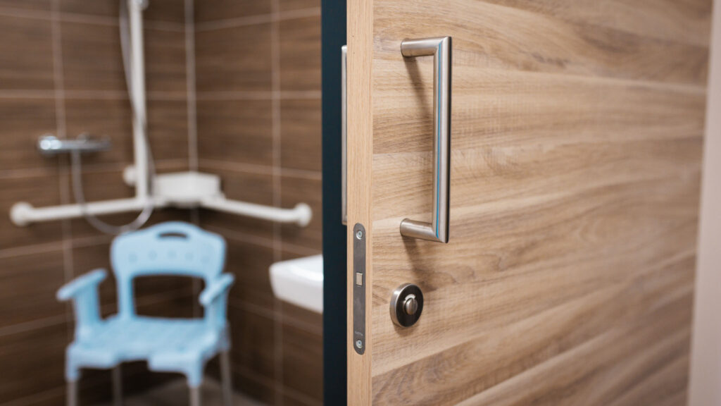 Detailaufnahme einer Holztür mit großem Edelstahlgriff. Im unscharfen Hintergrund ist ein barrierefreies Badezimmer mit einem blauen Duschstuhl zu erkennen.