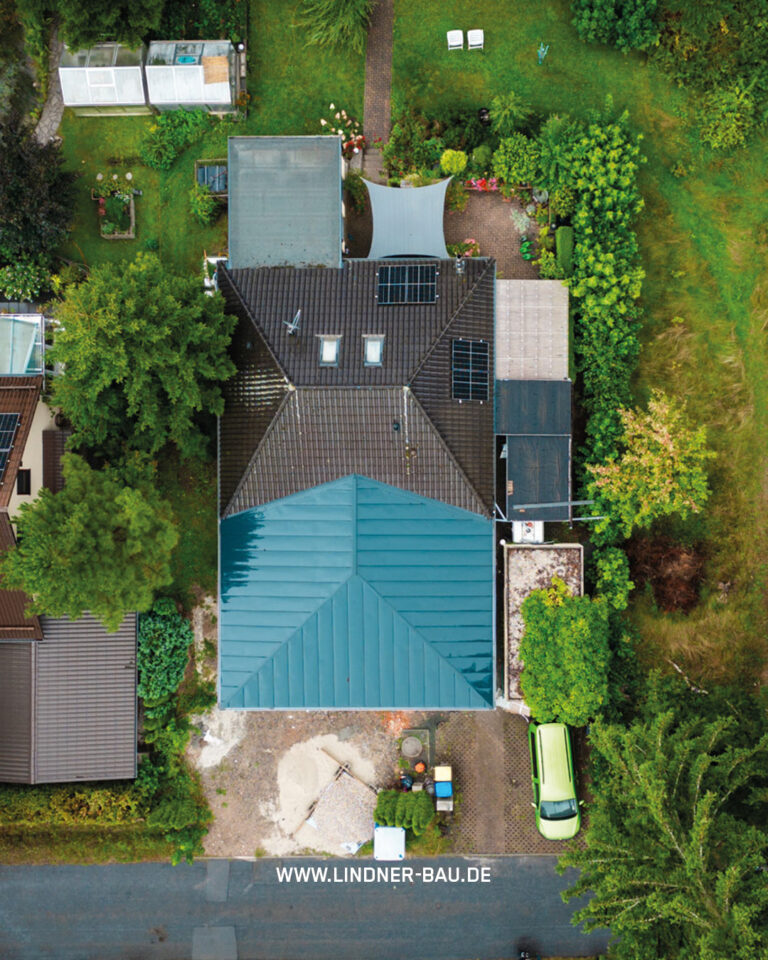 Dachsanierung und Carport-Neubau an einem Einfamilienhaus in einer Wohnsiedlung, aufgenommen aus der Vogelperspektive. Das Bild zeigt das dunkelbraune Ziegeldach mit installierten Solarpaneelen und das neue, große, türkisfarbene Trapezblechdach eines Carports oder einer Garage. Der Alt-Text enthält das Wasserzeichen und Keyword LINDNER-BAU.DE.