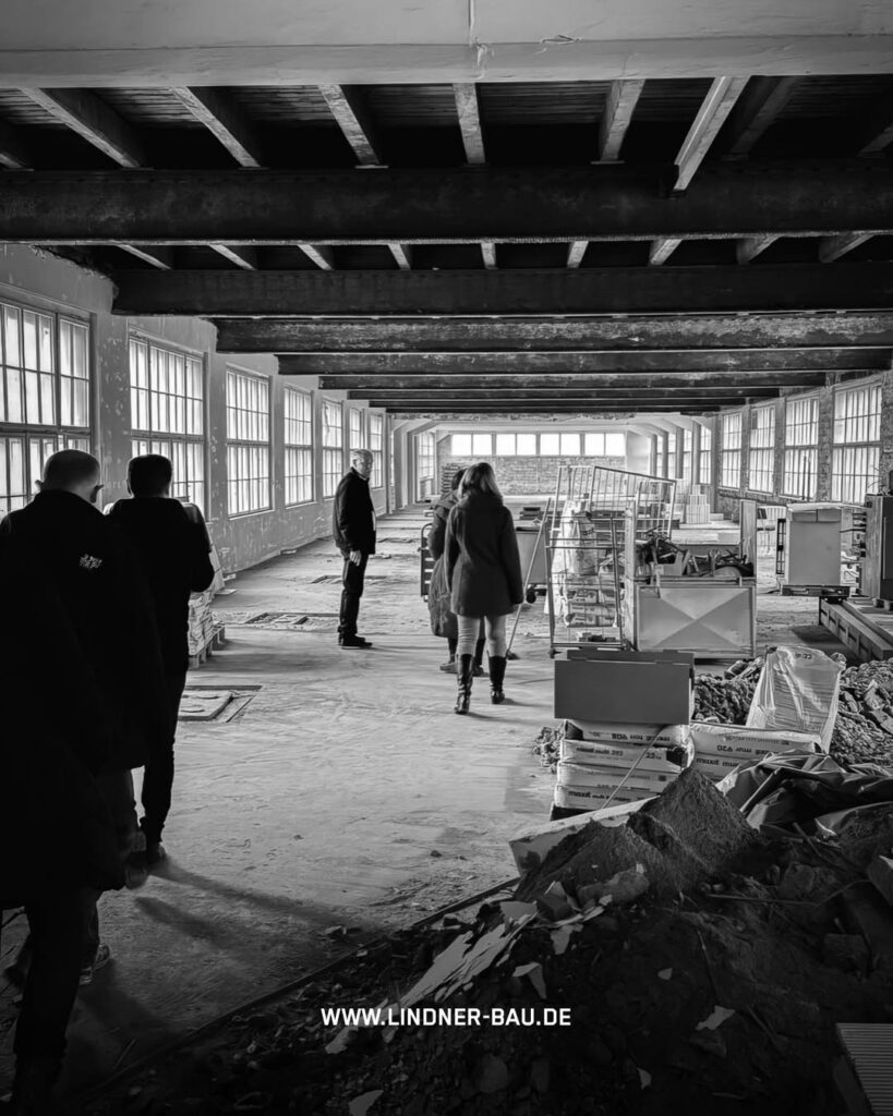 Schwarz-weiß-Foto des langen, hellen Fabrikflurs im Rogo-Werk mit Holzbalkendecke, vielen Fenstern und Bauschutt im Vordergrund, mit Personen bei einer Besichtigung.