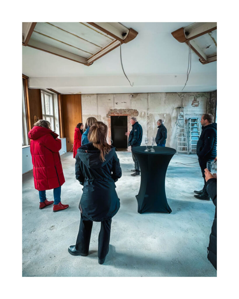 Besichtigung eines sanierungsbedürftigen, hellen Raumes im Rogo-Werk, in dem eine Gruppe von Menschen um einen Stehtisch mit Sektgläsern versammelt ist.