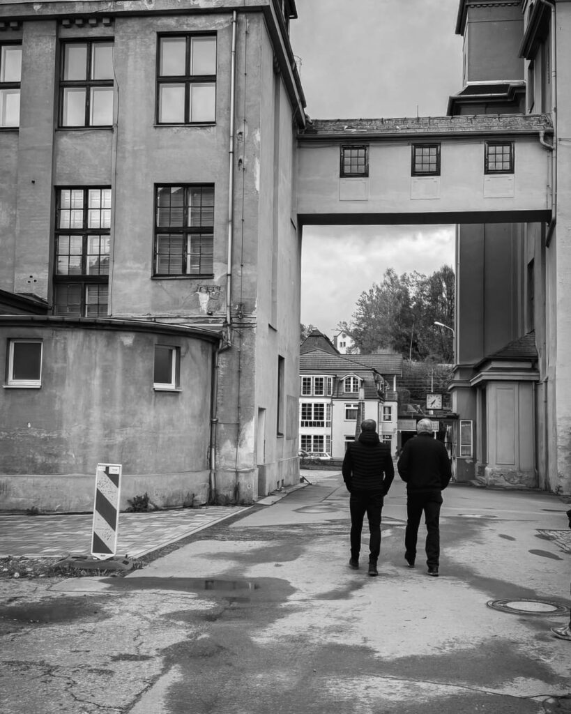 Schwarz-weiß-Aufnahme zweier Männer, die eine schmale Gasse im Rogo-Werk entlanggehen, vorbei an Fabrikgebäuden mit einem Verbindungssteg im oberen Bereich.