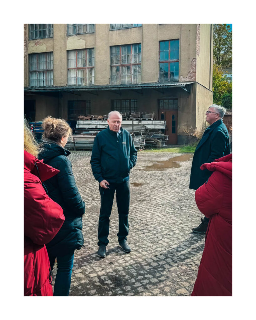 Eine Gruppe von Menschen, darunter ein Mann in dunkler Jacke, steht auf einem gepflasterten Innenhof vor einem alten Fabrikgebäude des Rogo-Werks, das zur Sanierung ansteht.
