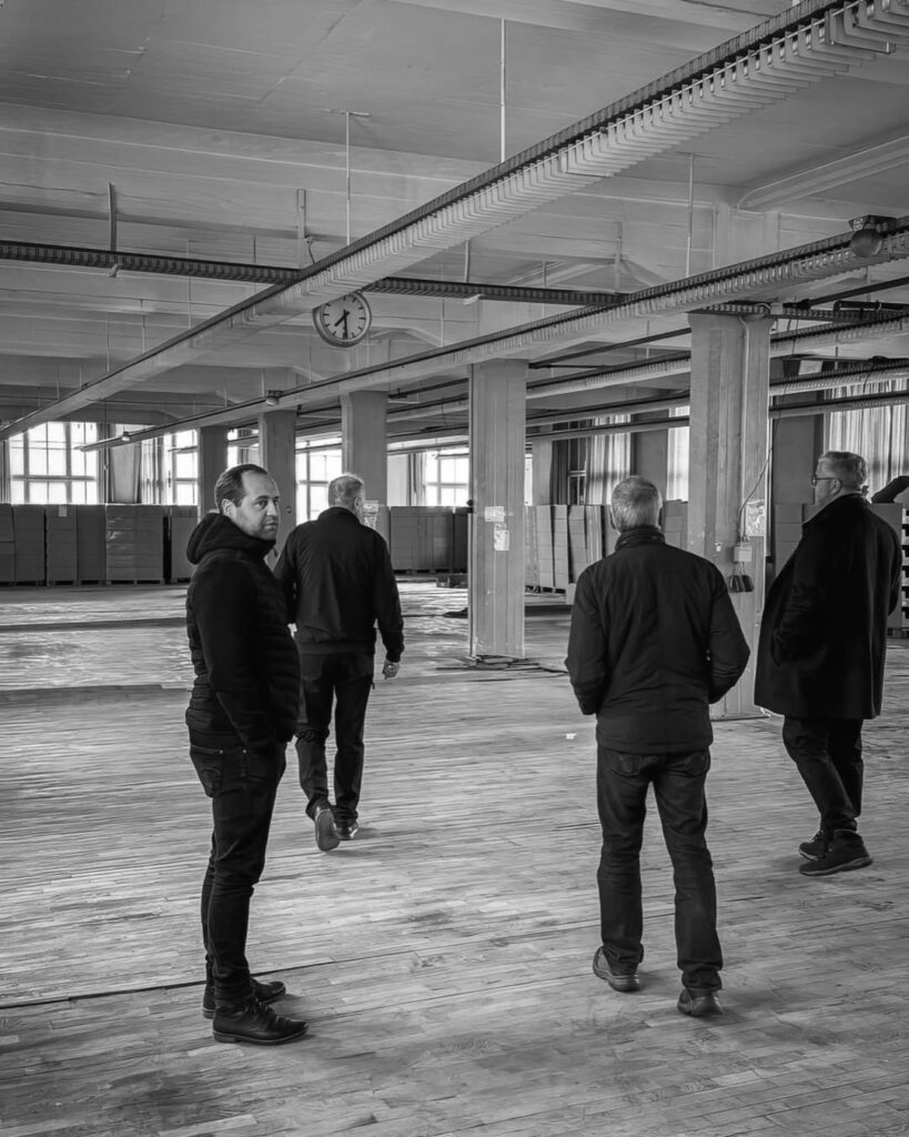 Schwarz-weiß-Bild einer Besichtigung in einer großen, leeren Fabrikhalle des Rogo-Werks mit Holzboden, Säulen, vielen Fenstern und einer Gruppe von vier Männern.