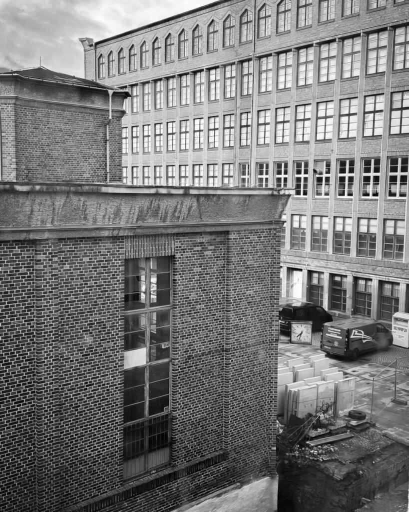 Schwarz-weiß-Ansicht des Rogo-Werks mit dem Blick von oben auf eine Backsteinmauer und das große, mehrstöckige Fabrikgebäude im Hintergrund mit vielen Fenstern.