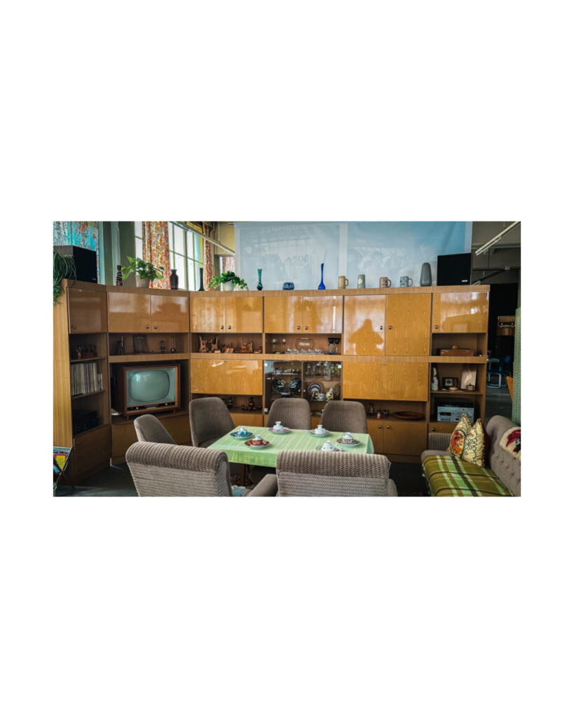 Innenansicht eines Raumes im Rogo-Werk, eingerichtet im Stil der DDR mit einer braunen Schrankwand, einem Tisch mit grünem Tuch und einem alten Röhrenfernseher.