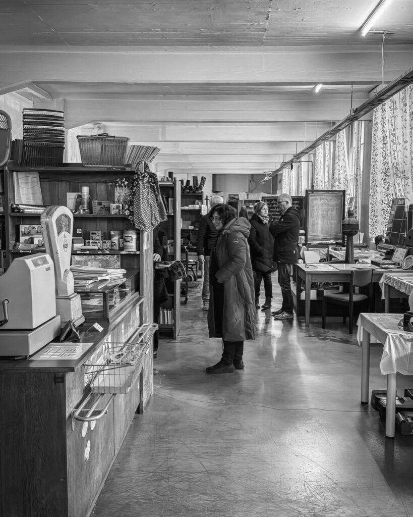 Schwarz-weiß-Foto eines temporären Ladens oder Archivs im Rogo-Werk mit alten Regalen, einer Kasse, Waage und einer Gruppe von Menschen im Hintergrund.