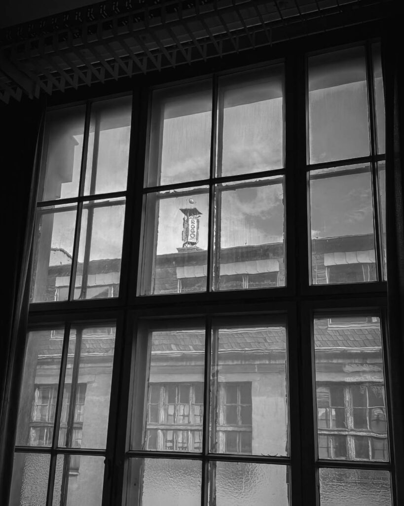 Schwarz-weiß-Aufnahme, die den Turm des Rogo-Werks mit dem Schriftzug "ROGO" durch ein großes, mehrteiliges Fenster im Inneren eines Fabrikgebäudes zeigt.