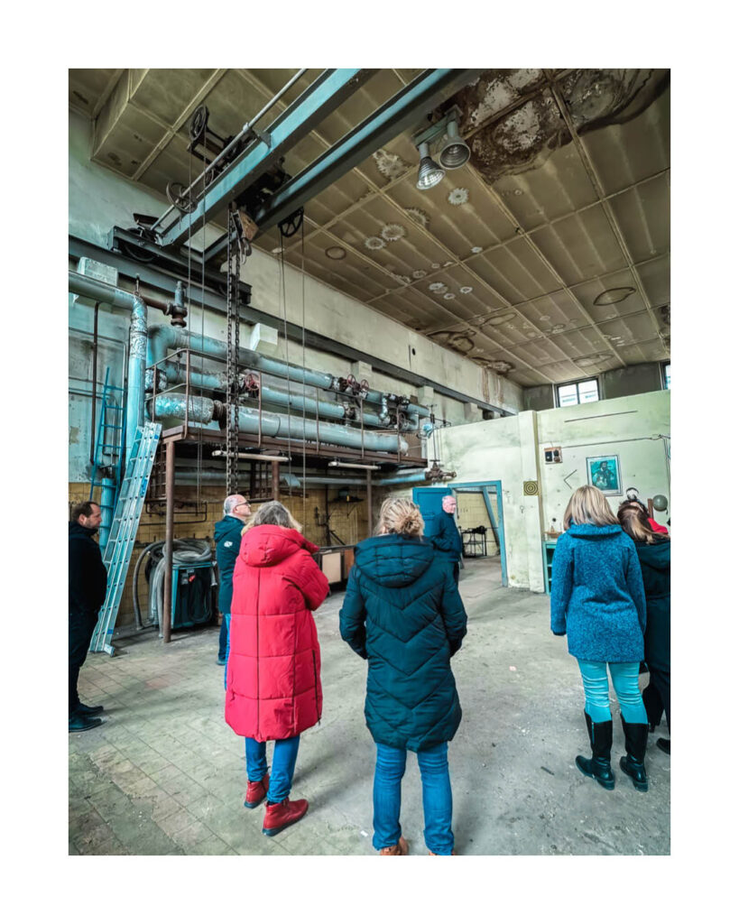 Besichtigung einer Industriehalle des Rogo-Werks mit einer Gruppe von Menschen, im Vordergrund große Rohre, Kettenzüge und eine Leiter.