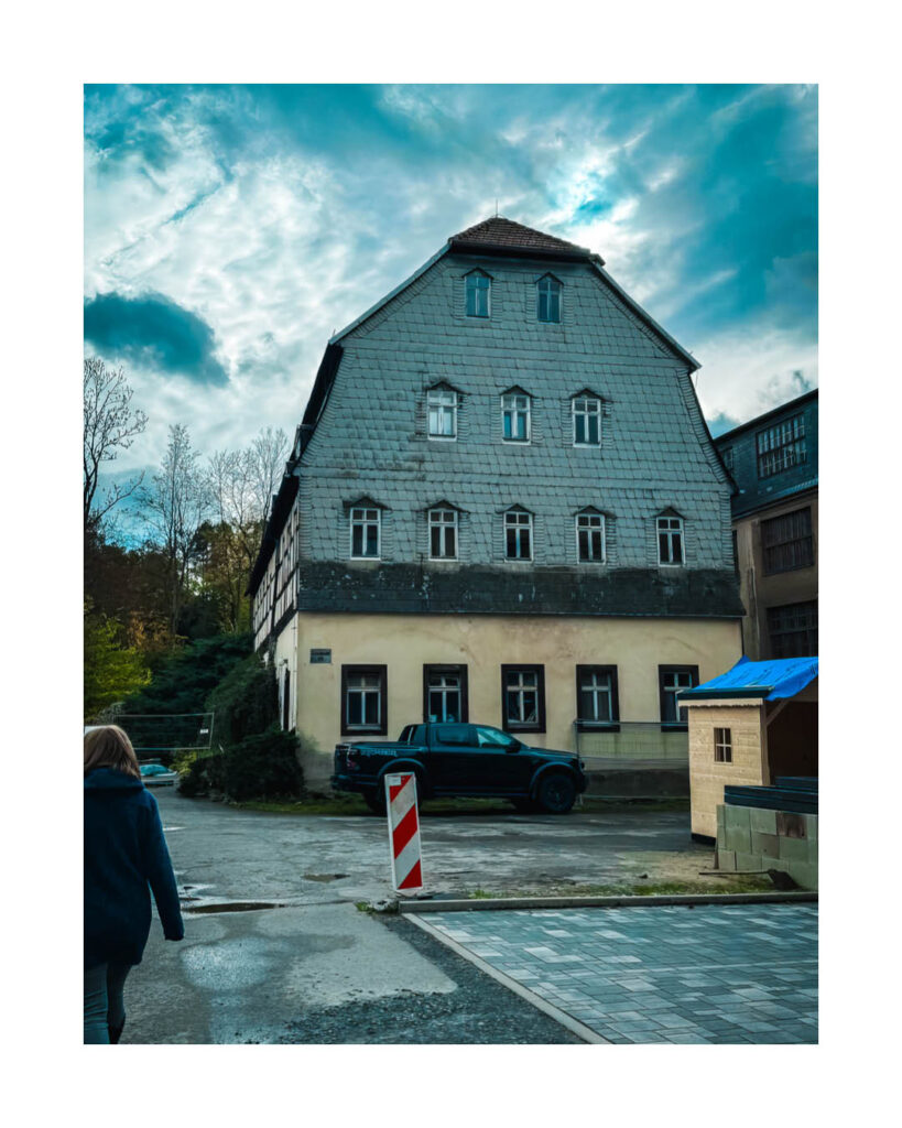 Historisches Gebäude mit hohem, schiefergedecktem Dach und weiß umrahmten Fenstern in der Umgebung des Rogo-Werks, mit dunklem Pickup-Truck davor.