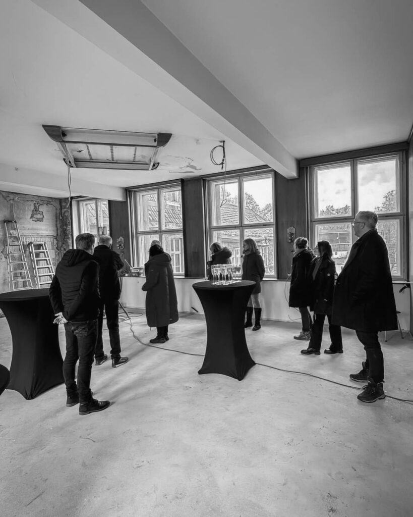 Schwarz-weiß-Aufnahme einer Gruppe von Menschen in einem großen, hellen Raum mit vielen Fenstern und zwei Stehtischen mit Sektgläsern während einer Besichtigung im Rogo-Werk.