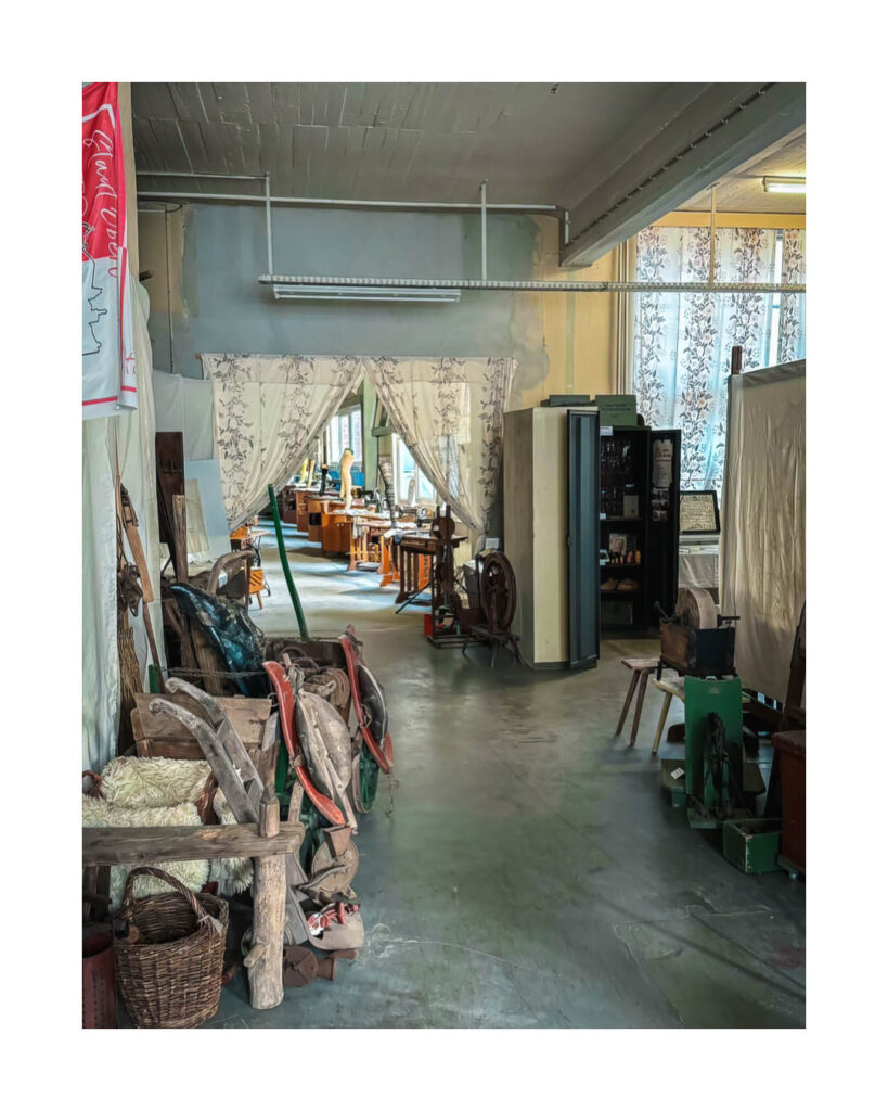 Werkstatt- oder Lagerraum des Rogo-Werks gefüllt mit alten Maschinen, Werkzeugen, Holzgegenständen und Textilien, mit einem Blick in einen hellen hinteren Bürobereich.