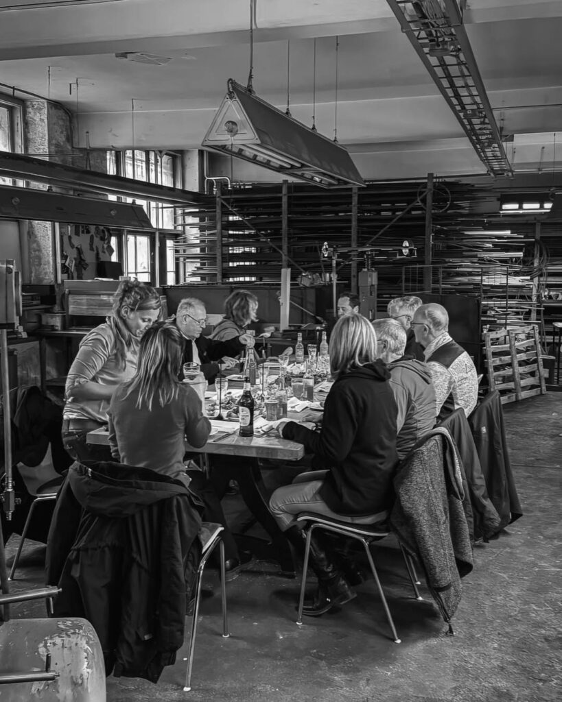 Schwarz-weiß-Foto einer Gruppe von Menschen, die in einer ehemaligen Industriehalle des Rogo-Werks an einem Tisch zusammensitzen und essen.