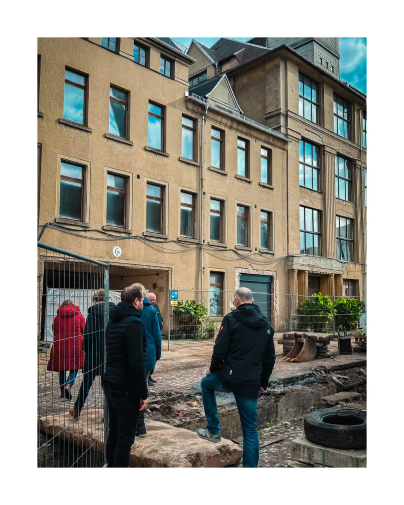 Gruppe von Menschen besichtigt den Baustellenbereich vor einem ockerfarbenen, mehrstöckigen Industriegebäude des ehemaligen Rogo-Werks.