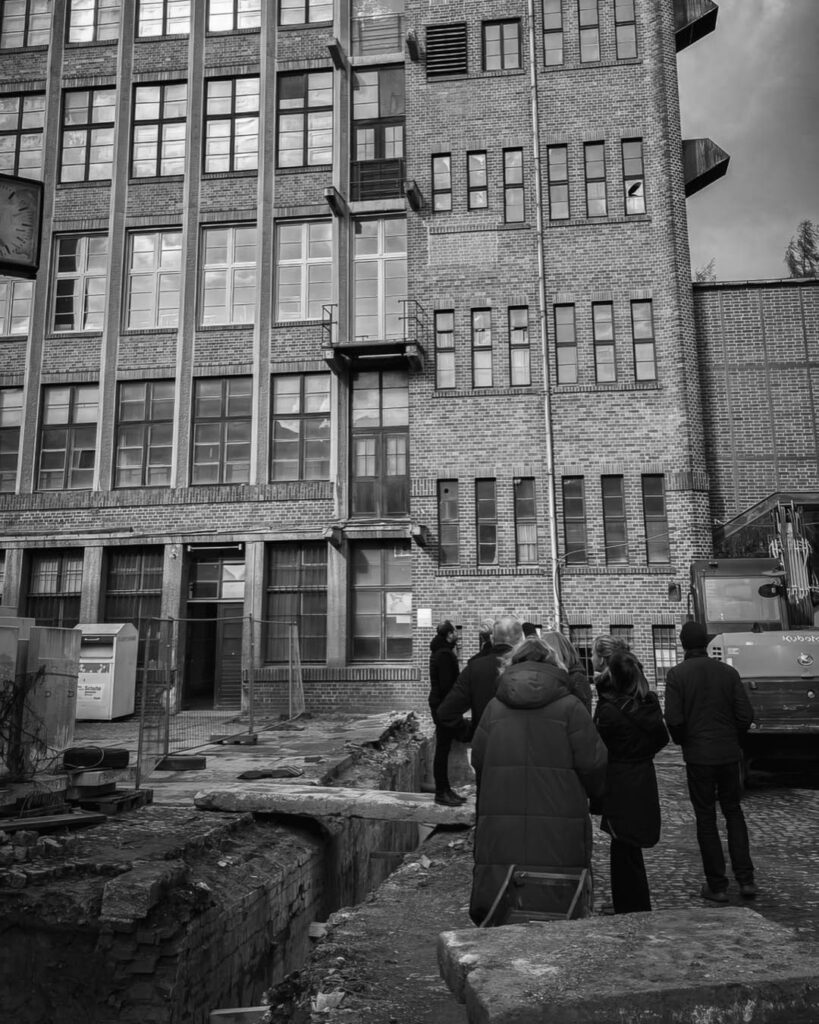 Schwarz-weiß-Foto einer Gruppe von Menschen an einer Baustelle vor der mehrstöckigen Backsteinfassade des Rogo-Werks, mit einem Bagger und einer Baugrube im Vordergrund.