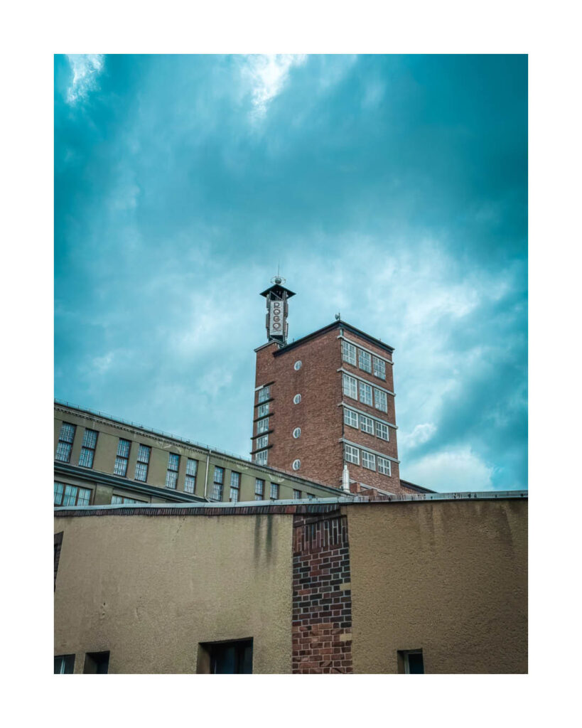 Außenansicht des markanten, turmartigen Backsteingebäudes des Rogo-Werks mit dem Schriftzug "ROGO" vor einem dramatischen, blauen Himmel.