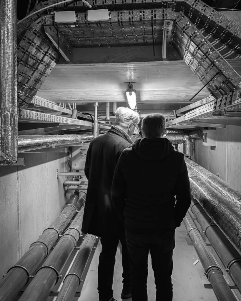 Schwarz-weiß-Foto von zwei Personen, die durch einen engen Technik- oder Rohrtunnel im Rogo-Werk mit vielen Leitungen und Kabeltrassen gehen.