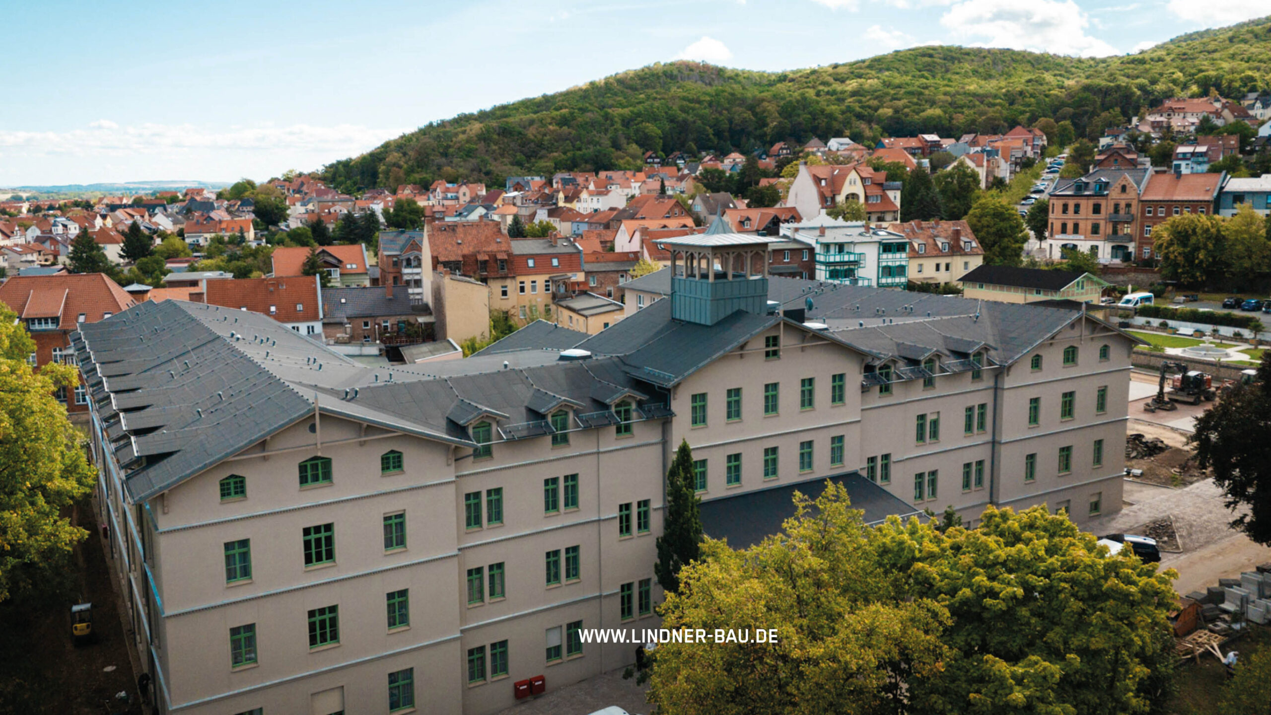 Luftaufnahme eines großen U-förmigen Gebäudes mit dunklem Dach und grünen Fensterrahmen in einer städtischen Umgebung, umgeben von kleineren Häusern und Bäumen.