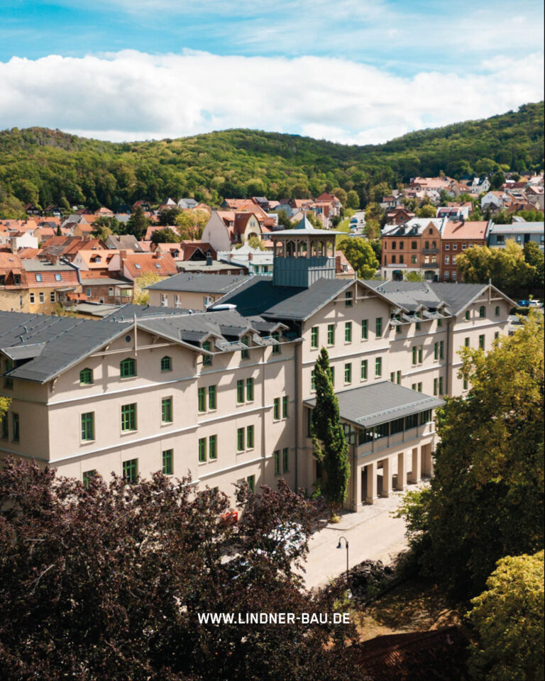 Luftaufnahme eines großen U-förmigen Gebäudes mit dunklem Dach und grünen Fensterrahmen in einer städtischen Umgebung, umgeben von kleineren Häusern und Bäumen.
