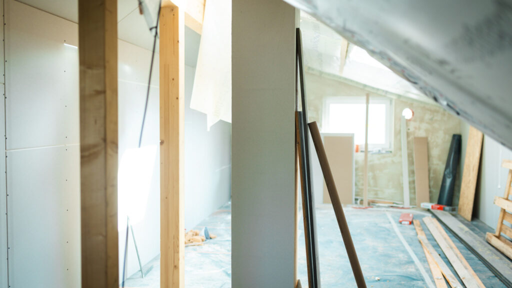Innenausbau eines Dachgeschosses mit Trockenbauwänden, Holzbalken und Schutzfolie am Boden.