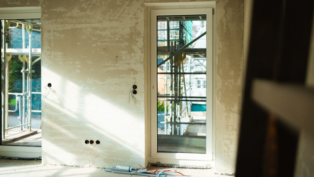 Innenansicht eines Neubaus mit frisch verputzten Wänden und einer bodentiefen Glastür mit Blick auf das Baugerüst.