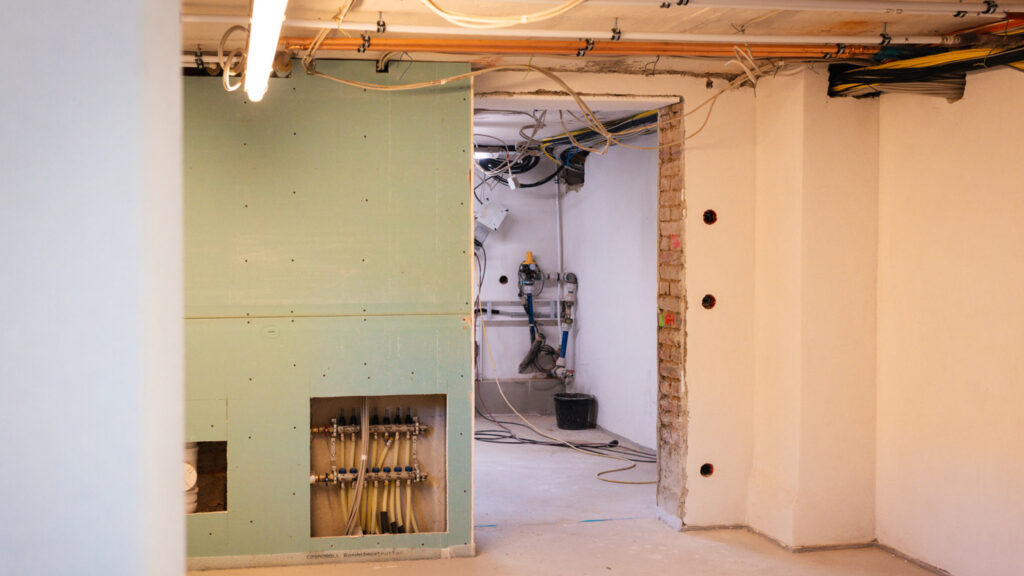 Innenansicht eines Raumes im Rohbau, mit freigelegter Installation für Fußbodenheizung (Verteiler) in einer Gipskartonwand und Blick in einen dahinterliegenden Technikraum mit Leitungen und Eimern.