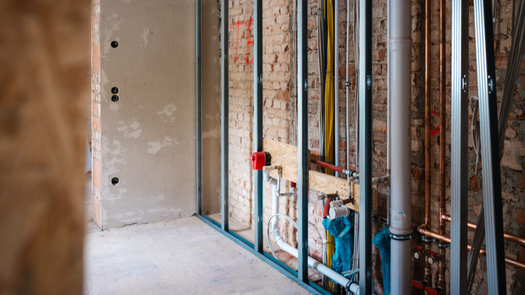 Nahaufnahme einer Trockenbauwand im Rohzustand. Sichtbar sind Metallständer, elektrische Leitungen und Sanitärrohre vor einer ursprünglichen Ziegelsteinwand.