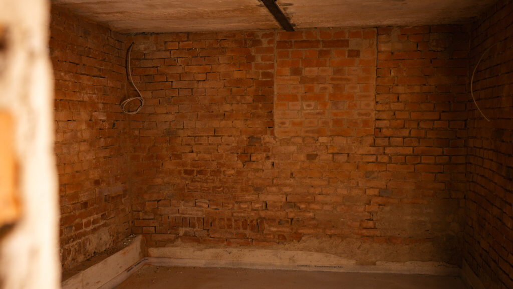 Ecke eines Raumes im Untergeschoss oder Keller mit freiliegender Backsteinwand und einem neu betonierten, niedrigen Sockelbereich am Boden.