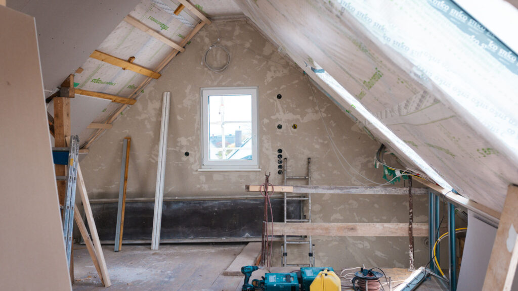 Dachgeschoss im Rohbau während der Sanierung. Man sieht ein dreieckiges Fenster in der Giebelwand, umgeben von Holzbalken, Dämmmaterial und neu montierten Gipskartonplatten.