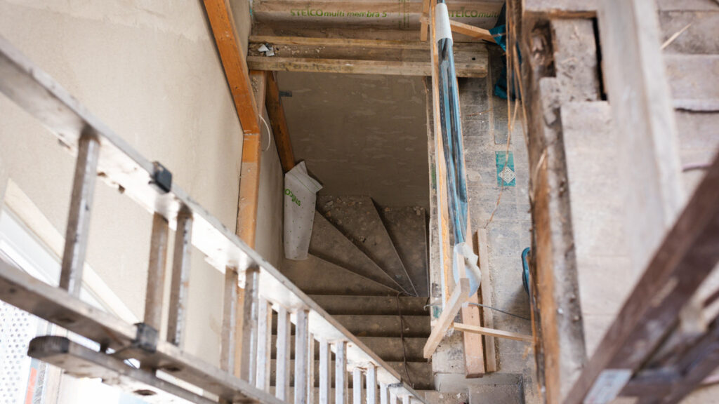 Blick von oben auf eine offene Treppe im Bau, die in das darunterliegende Geschoss führt. Eine Holzleiter und eine provisorische Absicherung im Vordergrund sind zu sehen.