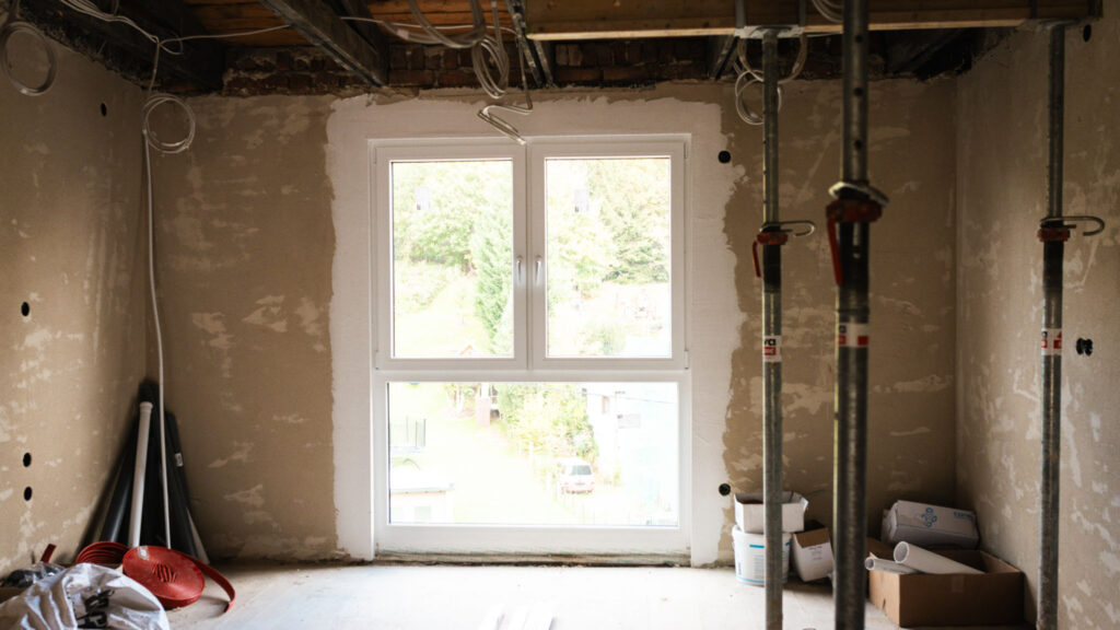 Innenansicht eines Zimmers im Rohbau mit einem neu eingebauten, großen Kunststoff-Doppelfenster. Stützpfeiler und Baumaterialien liegen im Raum.