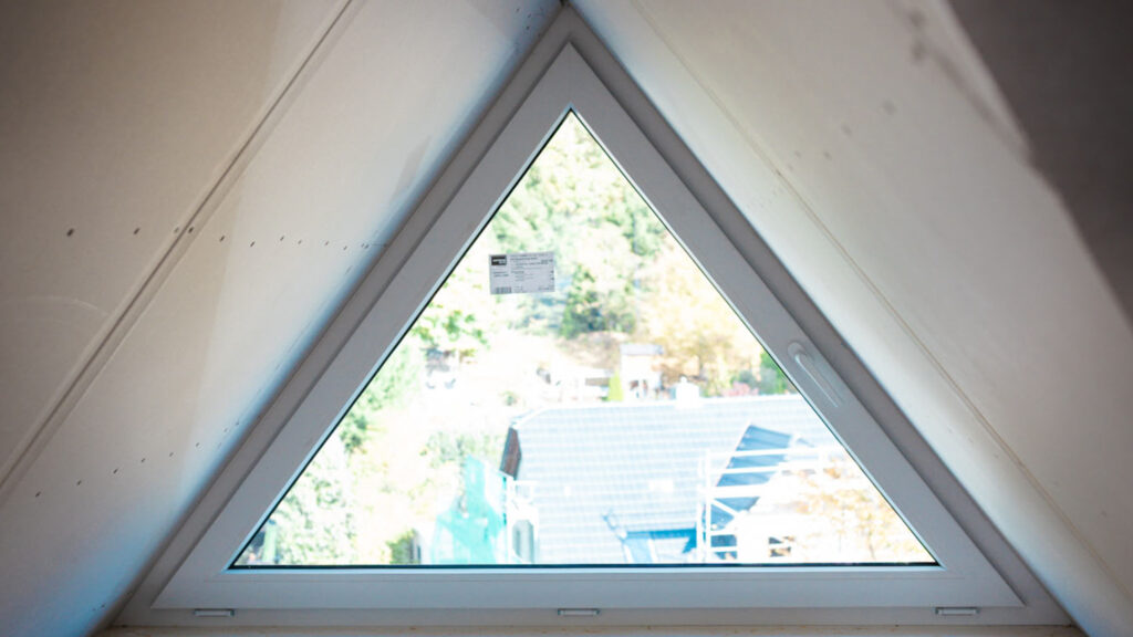 Nahaufnahme eines dreieckigen Giebelfensters im Dachgeschoss. Der Raum ist mit zugeschnittenen Gipskartonplatten verkleidet und befindet sich im frühen Stadium des Innenausbaus.