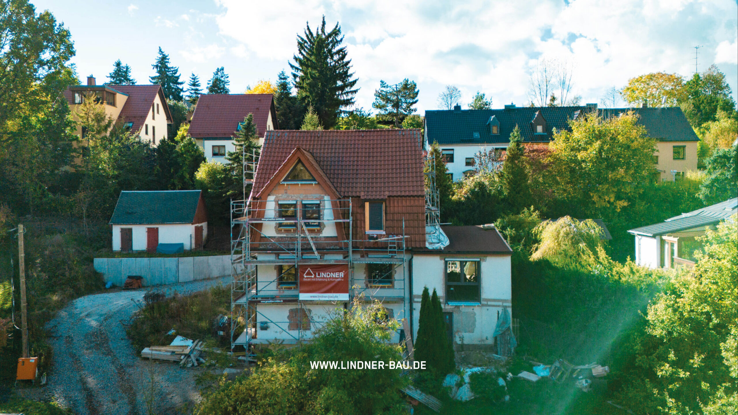 Außenaufnahme eines Einfamilienhauses während der Sanierung, umgeben von Gerüsten und einem roten Ziegeldach. Sichtbar ist ein "LINDNER"-Banner an der Fassade.