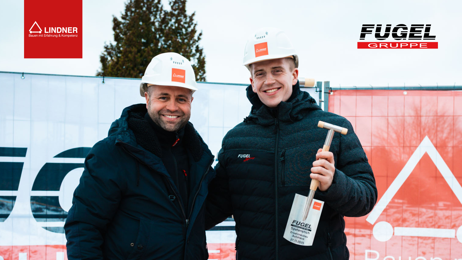 Spatenstich-Veranstaltung: Zwei Vertreter der Unternehmen Fugel Gruppe und Lindner Group, ausgestattet mit Schutzhelmen und warmer Kleidung, posieren mit einem Spaten beim offiziellen Baubeginn. Im Hintergrund sind die Logos der Lindner Group ("Bauen mit Erfahrung & Kompetenz") und der Fugel Gruppe zu sehen.