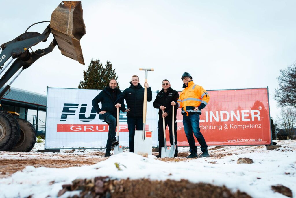 Niedrige Perspektive beim feierlichen Spatenstich der Fugel Gruppe und Lindner Group. Die vier Bauherren stehen mit Spaten vor der Werbeplane, wobei links im Hintergrund die Schaufel eines Baggers den Beginn der Bauarbeiten markiert.
