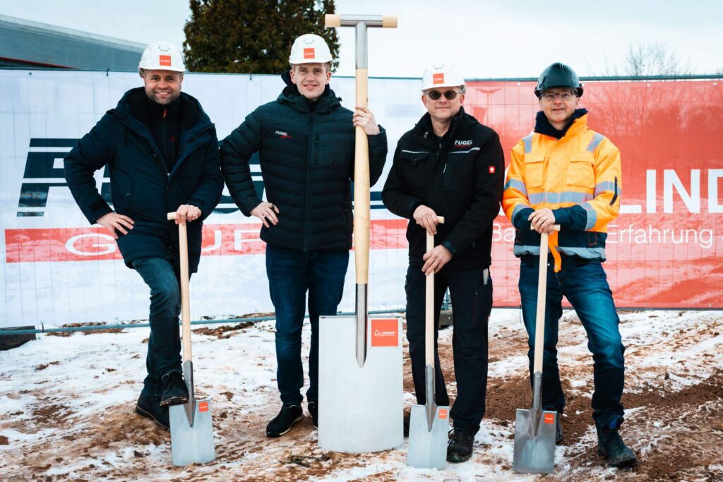 Gruppenfoto beim Spatenstich für den Ersatzneubau des Autohauses Fugel am 27.11.2025: Vier Vertreter der Fugel Gruppe und Lindner Group posieren in Bauhelmen und Winterkleidung mit symbolischen Spaten, darunter ein großer Dekorationsspaten, vor dem Firmenschild.