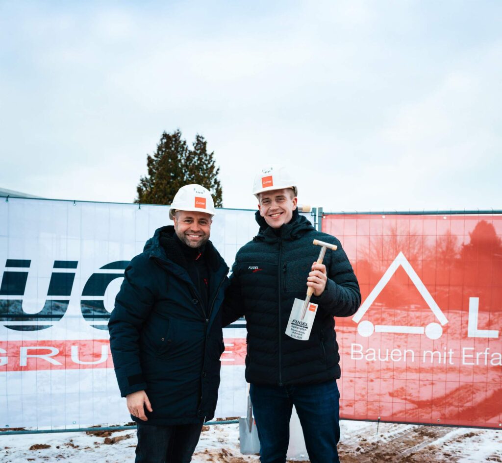 Zwei lächelnde Vertreter der Fugel Gruppe beim offiziellen Spatenstich für den Autohaus-Neubau. Der jüngere Mann hält den symbolischen Spaten mit den Veranstaltungsdetails, während sie vor der Baustellen-Plane mit den Logos der Fugel Gruppe und Lindner stehen.