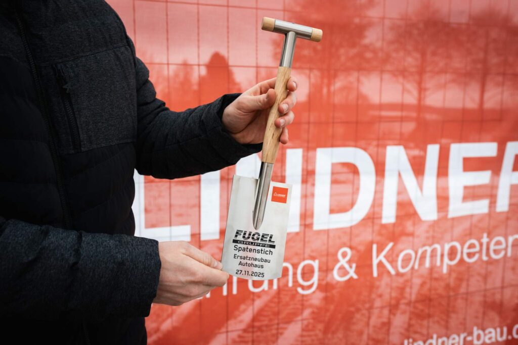 Nahaufnahme eines symbolischen Minispatens mit der Aufschrift: "FUGEL Spatenstich, Ersatzneubau Autohaus, 27.11.2025". Im unscharfen Hintergrund ist das rote Banner der Lindner Group mit dem Slogan "Bauen mit Erfahrung & Kompetenz" zu erkennen.