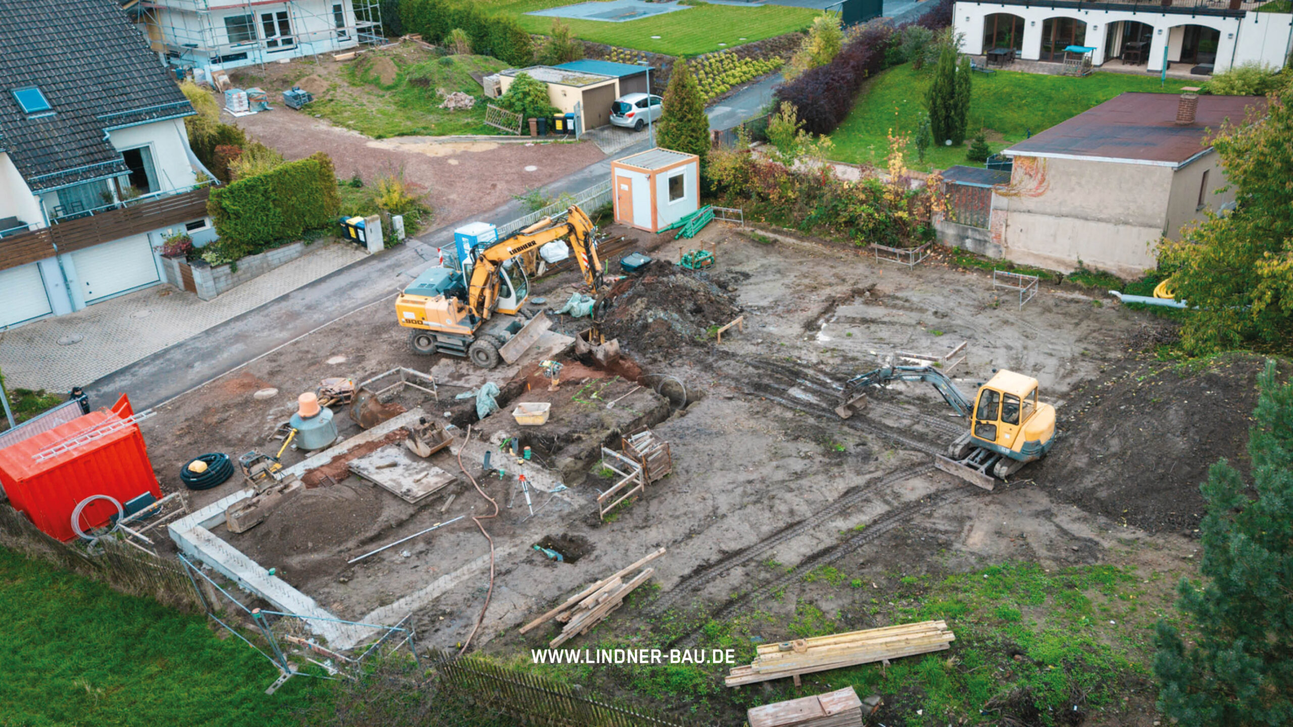 Weitwinkel-Luftaufnahme (Drohne) der gesamten Baustelle. Die Fundamentumrandung aus Beton ist zu erkennen. Zwei Bagger, ein roter Baucontainer und Baumaterial sind auf dem Bauplatz verteilt.