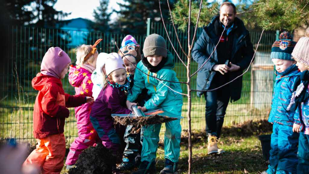 Eine Gruppe kleiner Kinder in bunter Winterkleidung hilft gemeinsam dabei, mit einer großen Schaufel Erde für eine Baumpflanzung zu bewegen, während der Chef der Baufirma Lindner lächelnd zuschaut.
