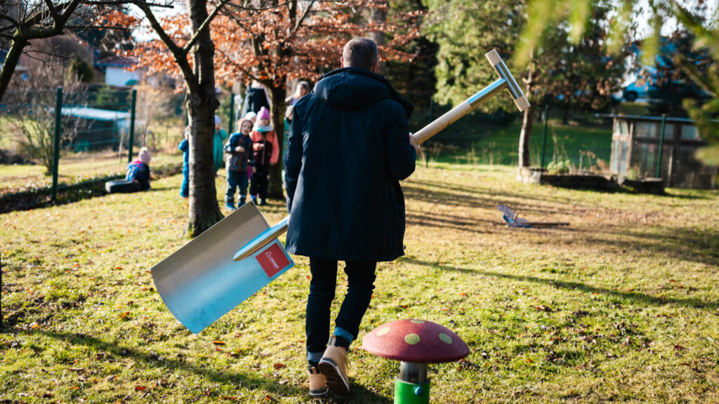 Rückansicht des Chefs der Baufirma Lindner in dunkler Winterjacke, der eine übergroße, glänzende Metallschaufel über eine sonnige Wiese in Richtung einer Gruppe von Kindern trägt.