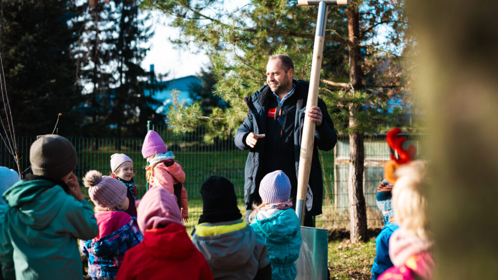 Der Chef der Baufirma Lindner erklärt einer Gruppe von Kindern im Freien die Baumpflanzung und hält dabei eine große Schaufel mit Lindner-Logo fest.