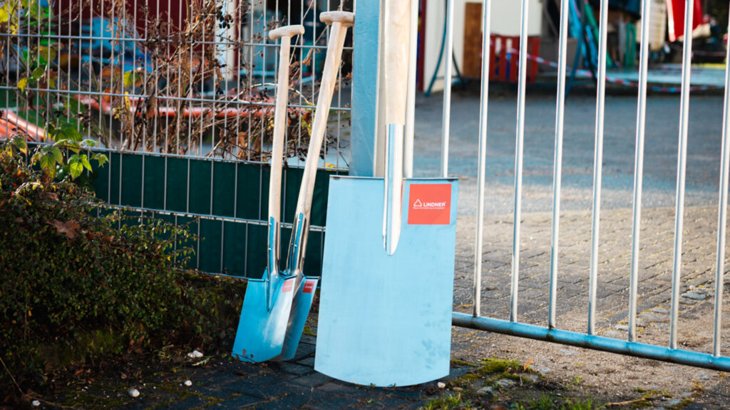 Drei hochwertige Spaten aus Edelstahl mit Holzstielen und rotem Lindner-Logo lehnen ordentlich an einem Metallzaun im Außenbereich.