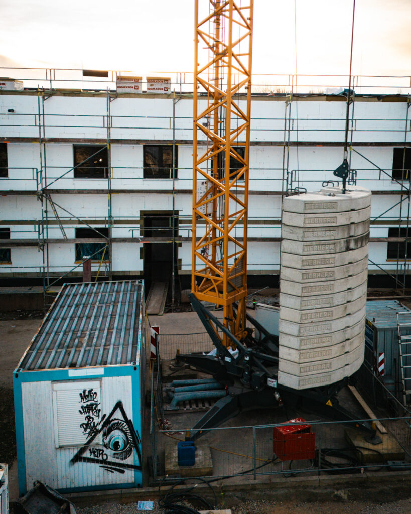 Nahaufnahme eines Liebherr Turmdrehkrans mit Gegengewichten vor der eingerüsteten Fassade eines Neubaus.