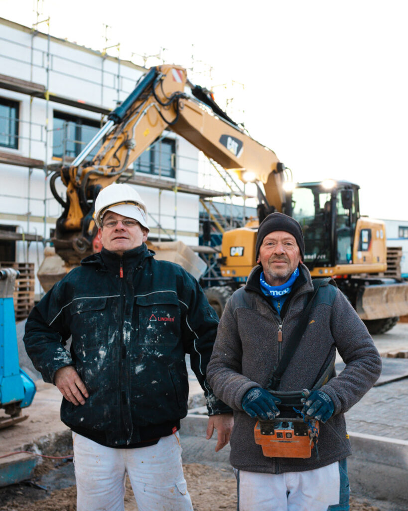 Zwei erfahrene Bauleiter von Lindner Bau in Arbeitskleidung vor einem CAT-Bagger auf einer Baustelle.